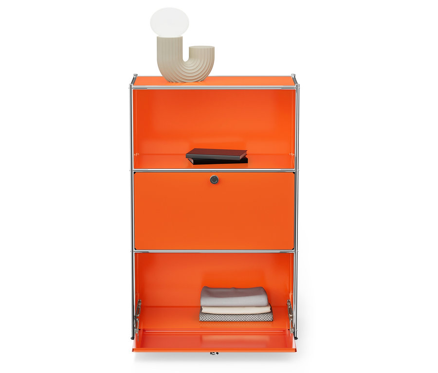 Narancssárga szekrény három rekesszel és ezüst kerettel. A felső polcon egy világos bézs lámpa és egy fekete könyv található. Az alsó rekesz nyitva van, és összehajtott anyagokat tartalmaz.