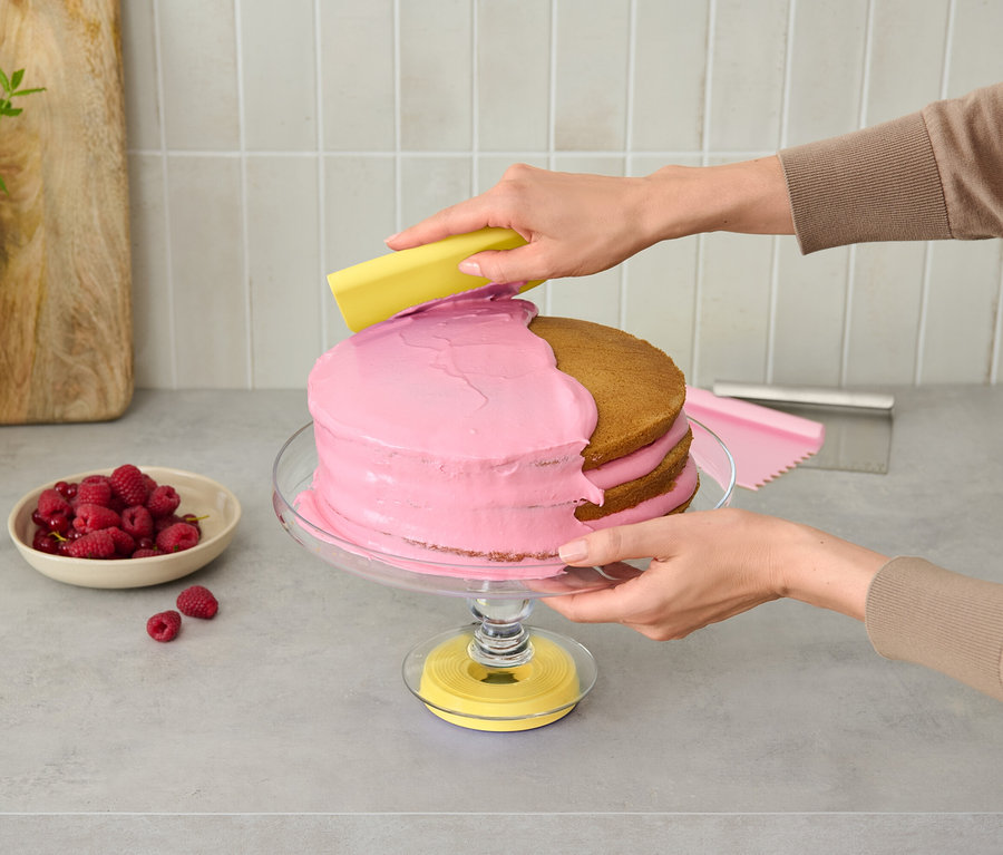 Egy személy rózsaszín tortát díszít egy üvegállványon sárga eszközzel.