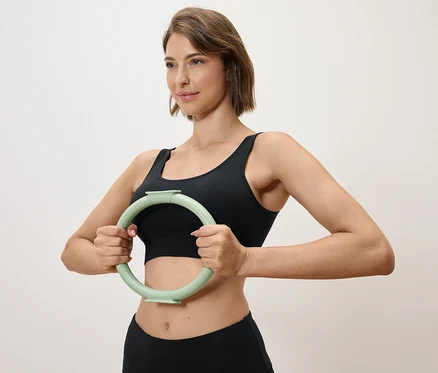 Nő tart maga előtt egy világoszöld edzőkört, fekete sportfelsőt és leggingset visel.