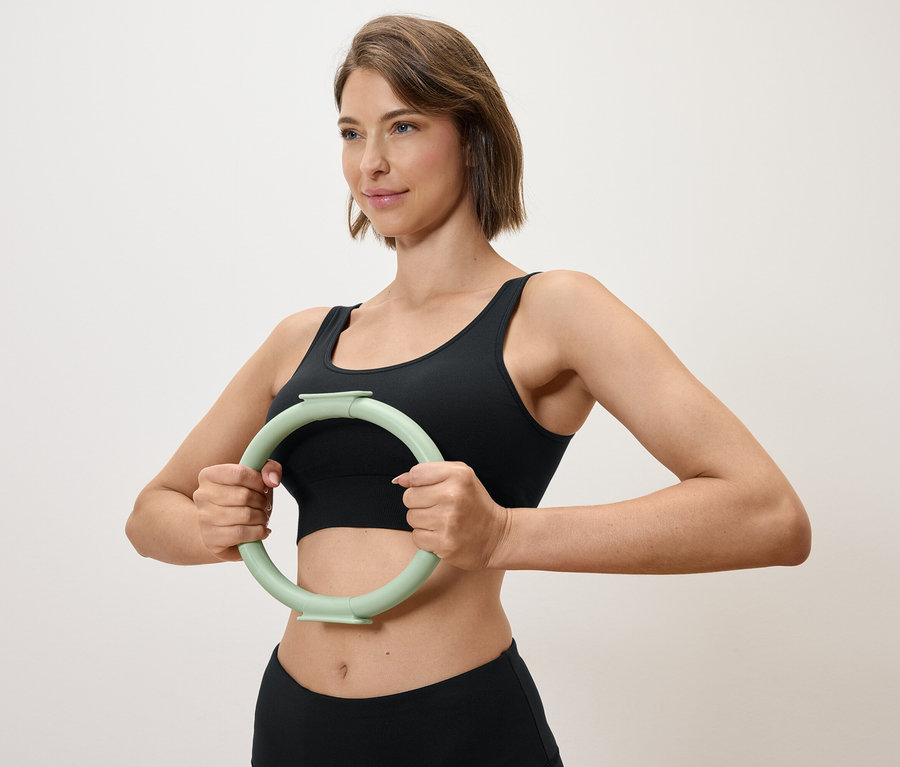 Nő tart maga előtt egy világoszöld edzőkört, fekete sportfelsőt és leggingset visel.
