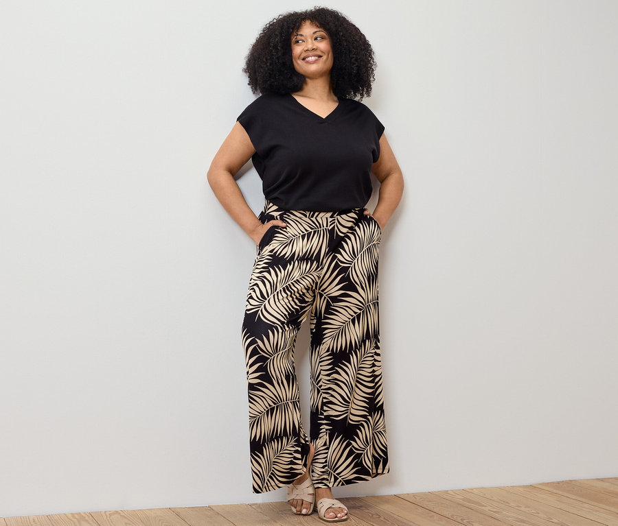 Nő pózol fekete, rövid ujjú, finom kötésű pulóverben és nyomott mintás palazzo nadrágban.