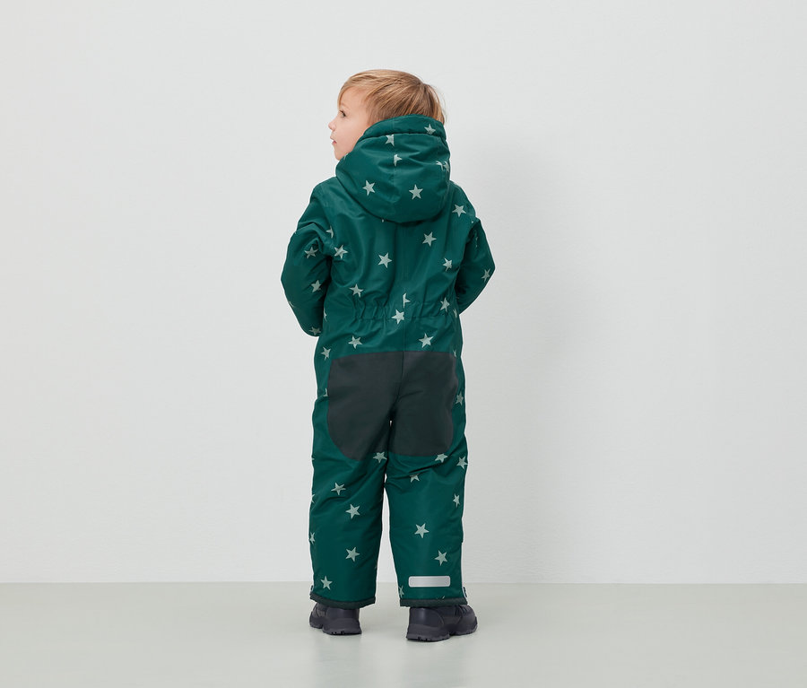 Egy fiú zöld, csillagmintás overallban áll háttal a kamerának.