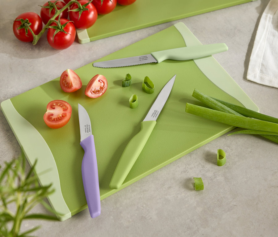 Zöld vágódeszka szeletelt paradicsommal, újhagymával és három, színes nyelű késsel.