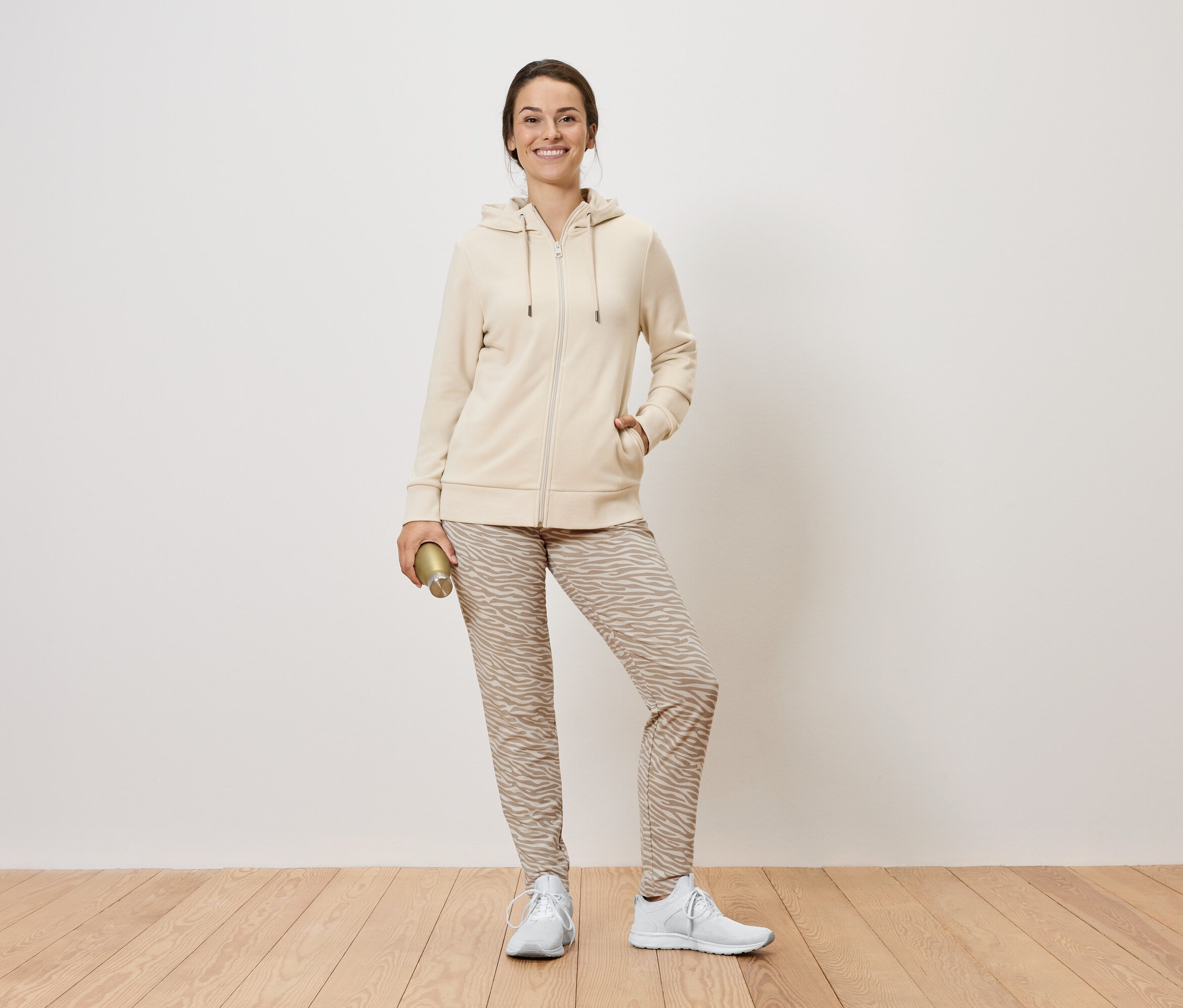 Egy nő áll és egy arany színű kulacsot tart. Bézs színű, cipzáros kapucnis pulóvert, mintás nadrágot és fehér tornacipőt visel.