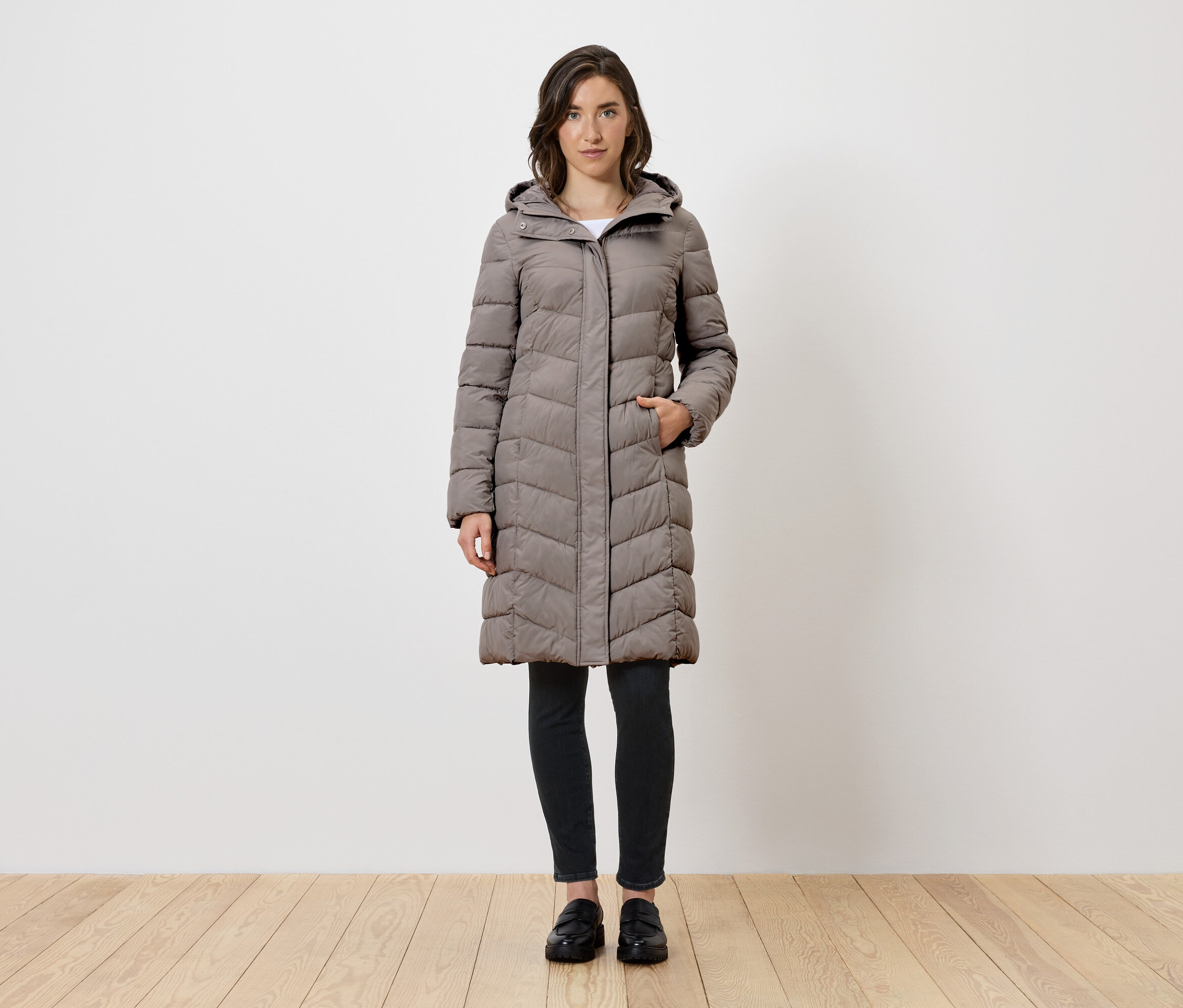 Egy nő áll, hosszú szürke, kapucnis pehelykabátot, fekete leggingst és fekete cipőt visel.
