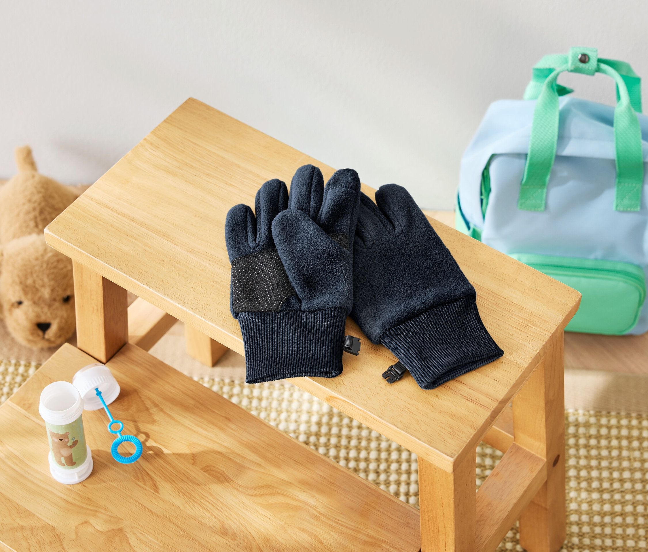 A fa zsámolyon egy pár sötétkék kesztyű fekszik, mellette egy buborékfújó és egy plüsskutya. A háttérben egy világoskék hátizsák látható mentazöld részletekkel.