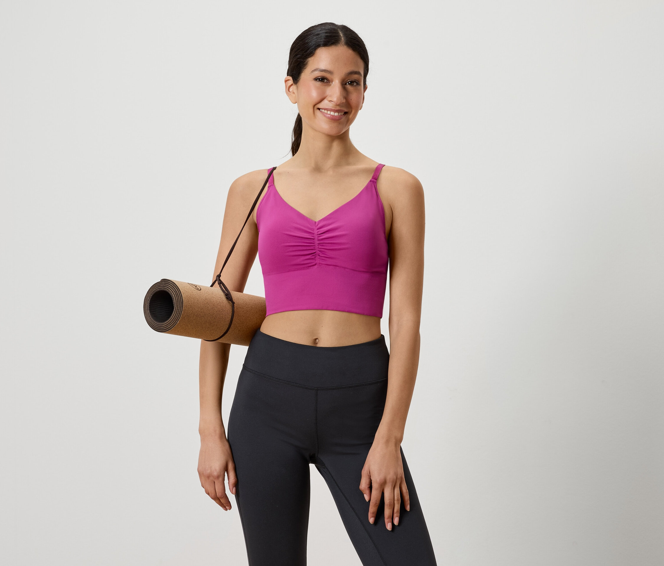 Nő barna jógaszőnyeggel a vállán, rózsaszín sportfelsőben és fekete leggingsben.
