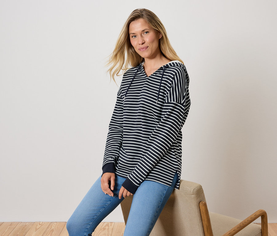 Egy nő ül egy széken, kék farmerben és egy csíkos kapucnis pulóverben.