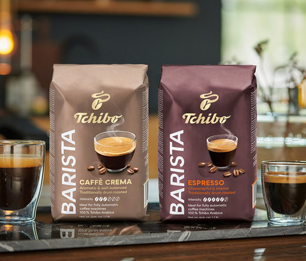 Két pohár kávé és két csomag Tchibo BARISTA Caffè Crema szemes kávé áll egy márványlapon.