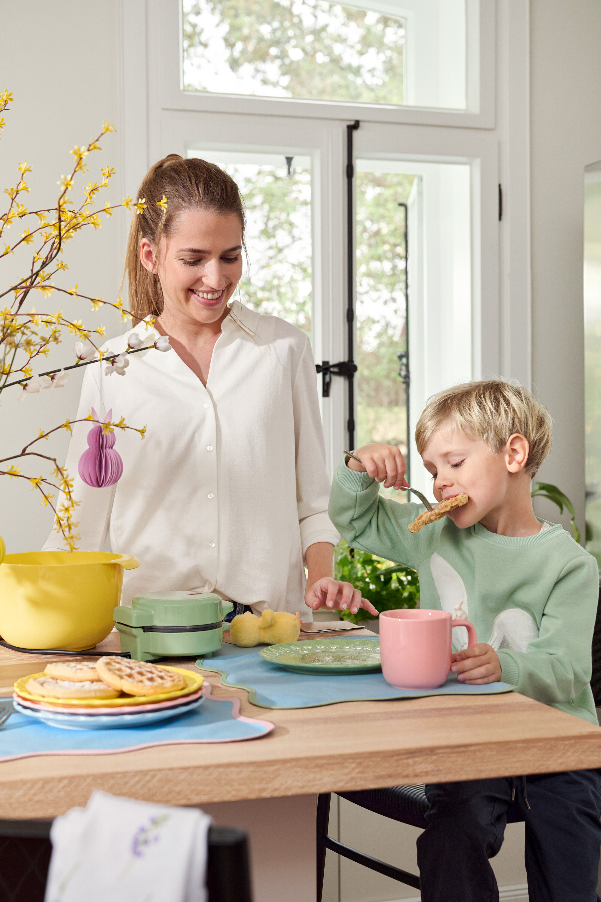 Nő és gyerek az asztalnál. A gyerek villával eszi a gofrit, a nő mosolyog.