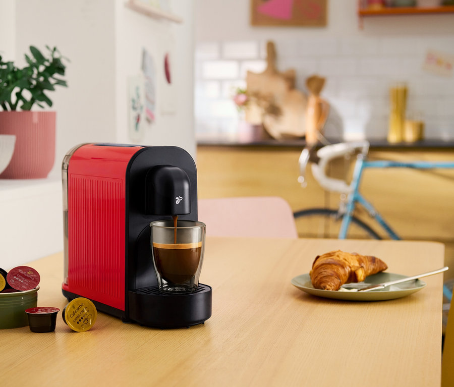 Egy piros kapszulás kávéfőző kávéval tölt egy csészét. Az asztalon egy croissant, egy tál kávékapszula és egy cserepes növény található.
