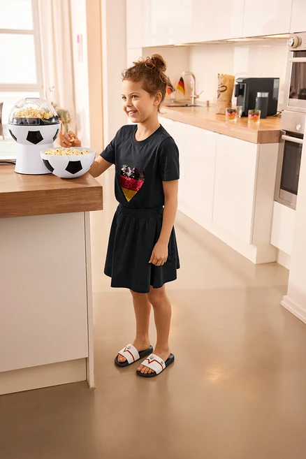 Egy lány pattogatott kukoricát eszik a foci pattogatott kukorica gép mellett. Kinder-Jerseyrock mit Hose, Kinder-T-Shirt mit Wendepailletten, Herz és Badelatschen van rajta.