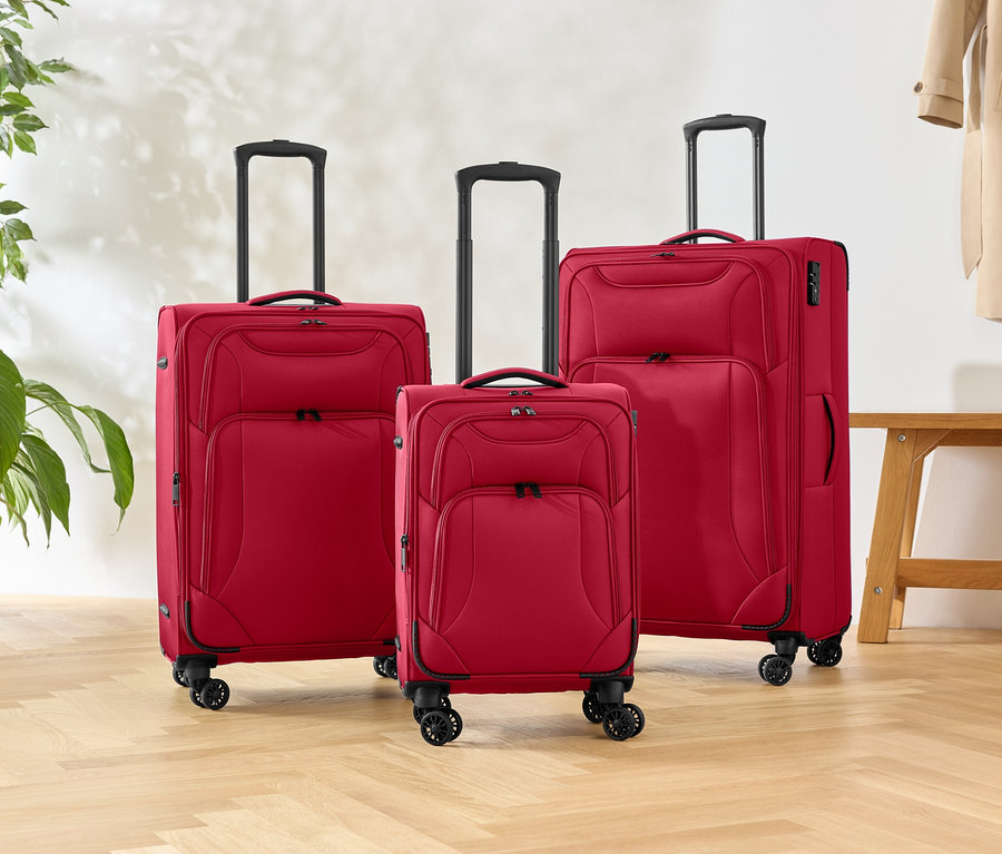 Három különböző méretű piros gurulós bőrönd áll egy fapadlón, mellettük egy növény és egy pad.