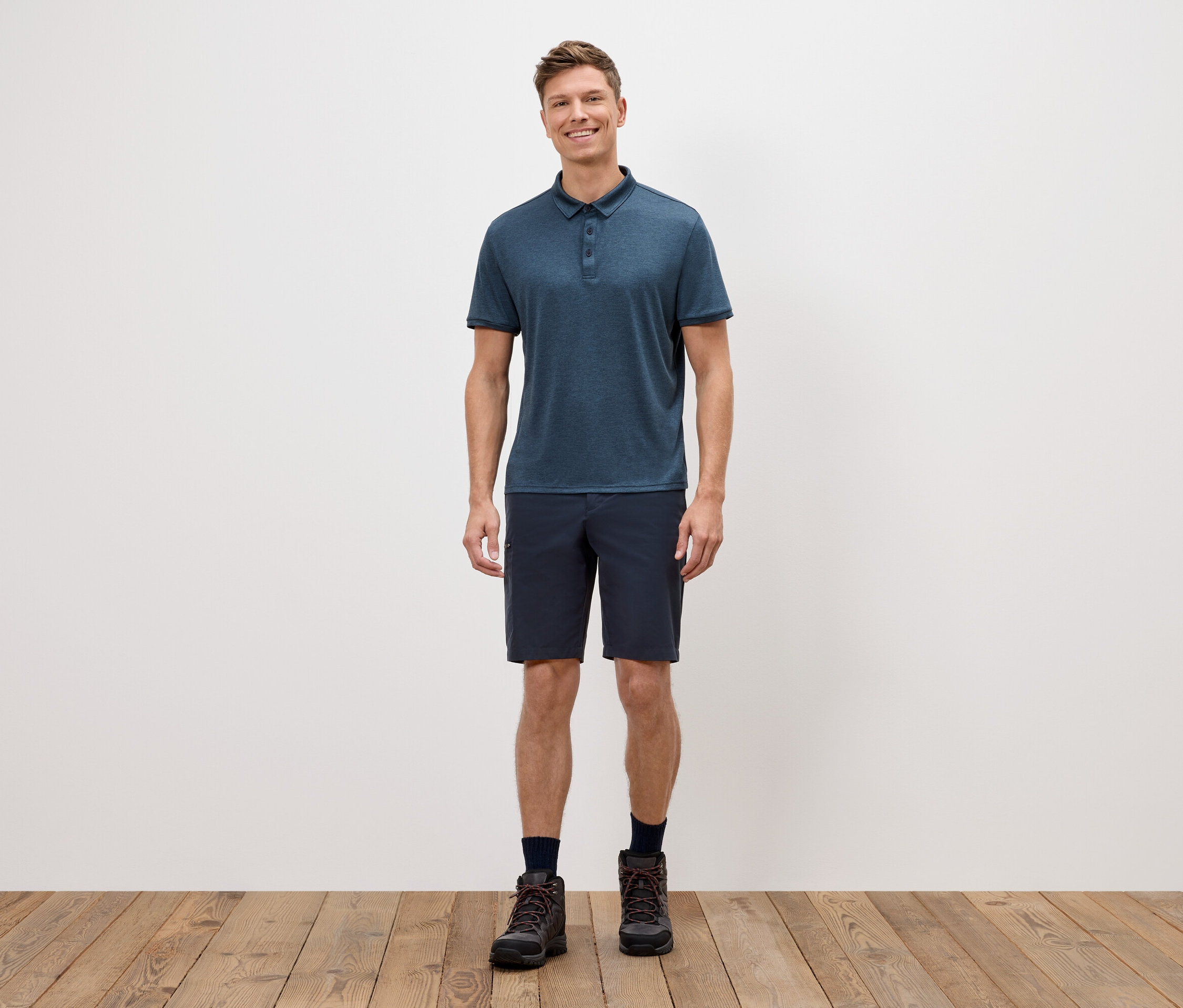 Egy férfi áll egy fehér fal előtt, kék pólót, rövidnadrágot és túrabakancsot visel.