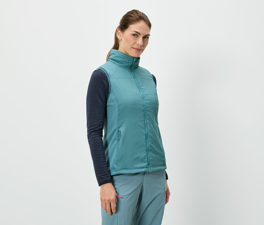 Egy nő áll, és kék mellényt visel egy hosszú ujjú felső fölött, kék nadrággal kombinálva.