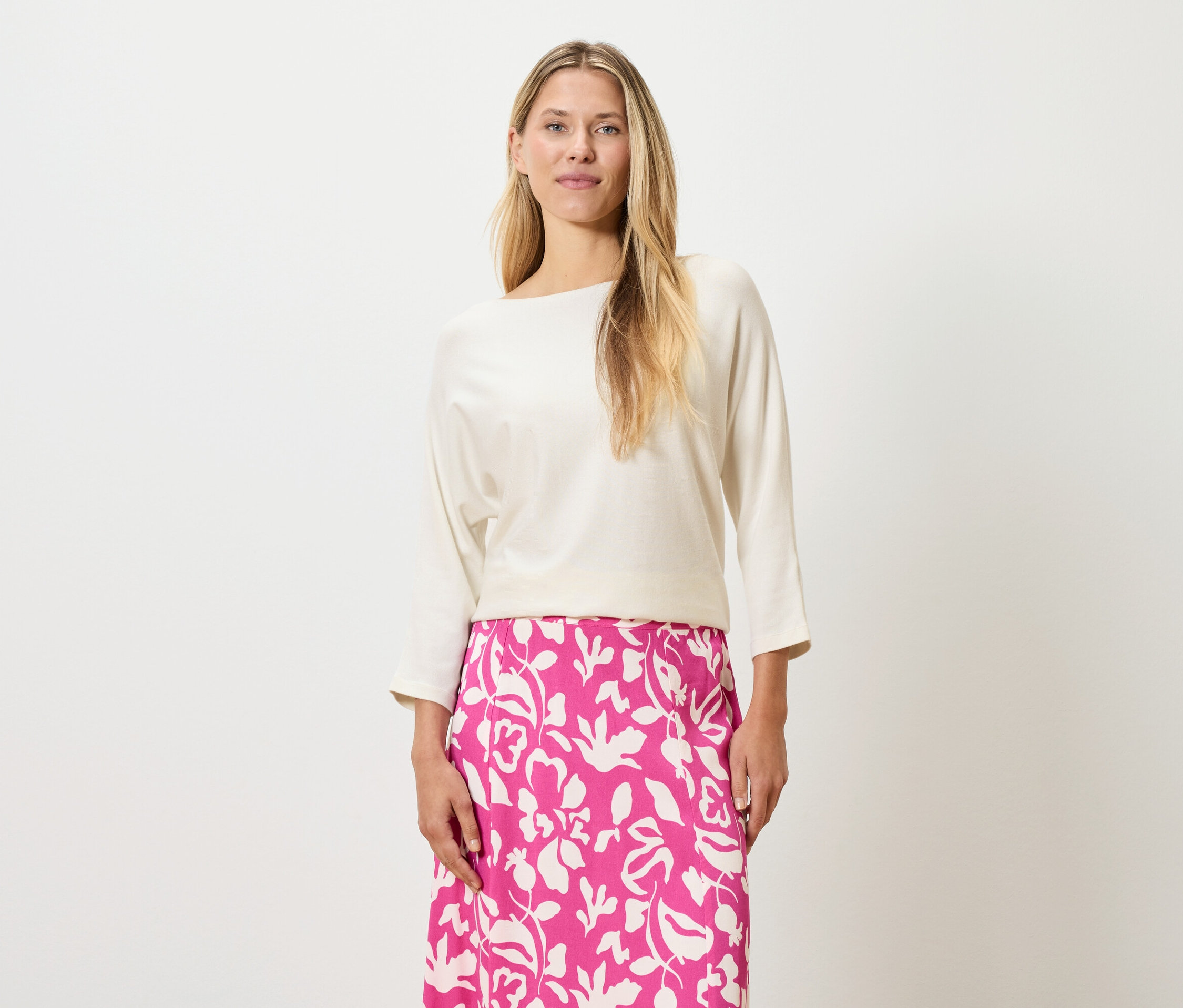 Egy szőke hajú nő krémszínű felsőt és rózsaszín, virágmintás szoknyát visel.