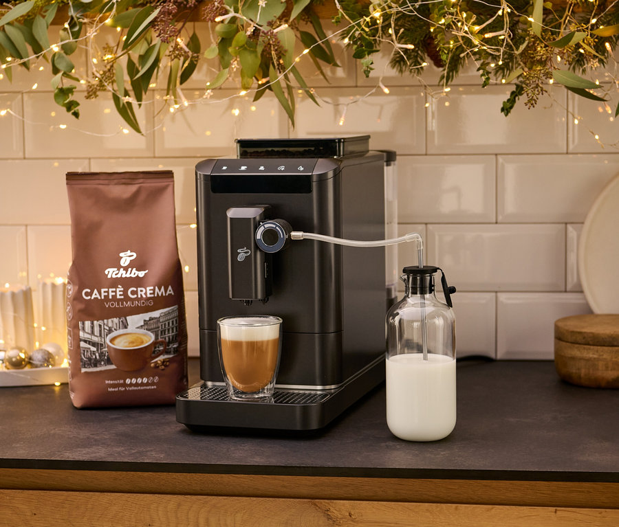 Egy »Esperto 2 Milk« Granite Black kávéfőző egy csésze kávéval és egy csomag Crema kávéval. A tejet egy üvegedényben tárolják.