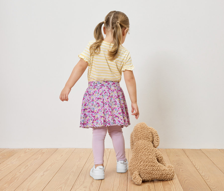 Hátulról látható egy copfos kislány sárga-fehér csíkos felsőben, virágos szoknyában, leggingsben és tornacipőben, egy plüssmackó mellett.