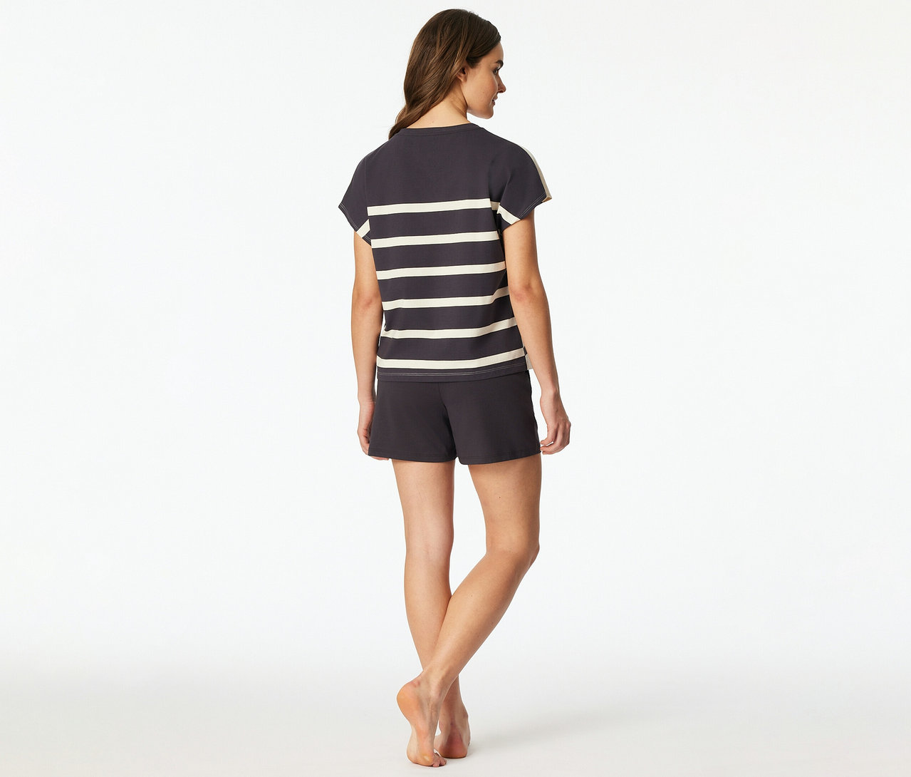 Egy nő háttal áll a nézőnek, sötétszürke SCHIESSER rövidnadrágos pizsamát visel csíkos felsővel és hozzá illő rövidnadrággal. Mezítláb van.
