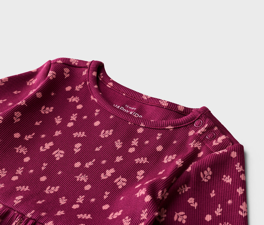Egy bordó, bordázott, hosszú ujjú póló közeli képe, világos rózsaszín virágmintával. A vállán két patent látható.