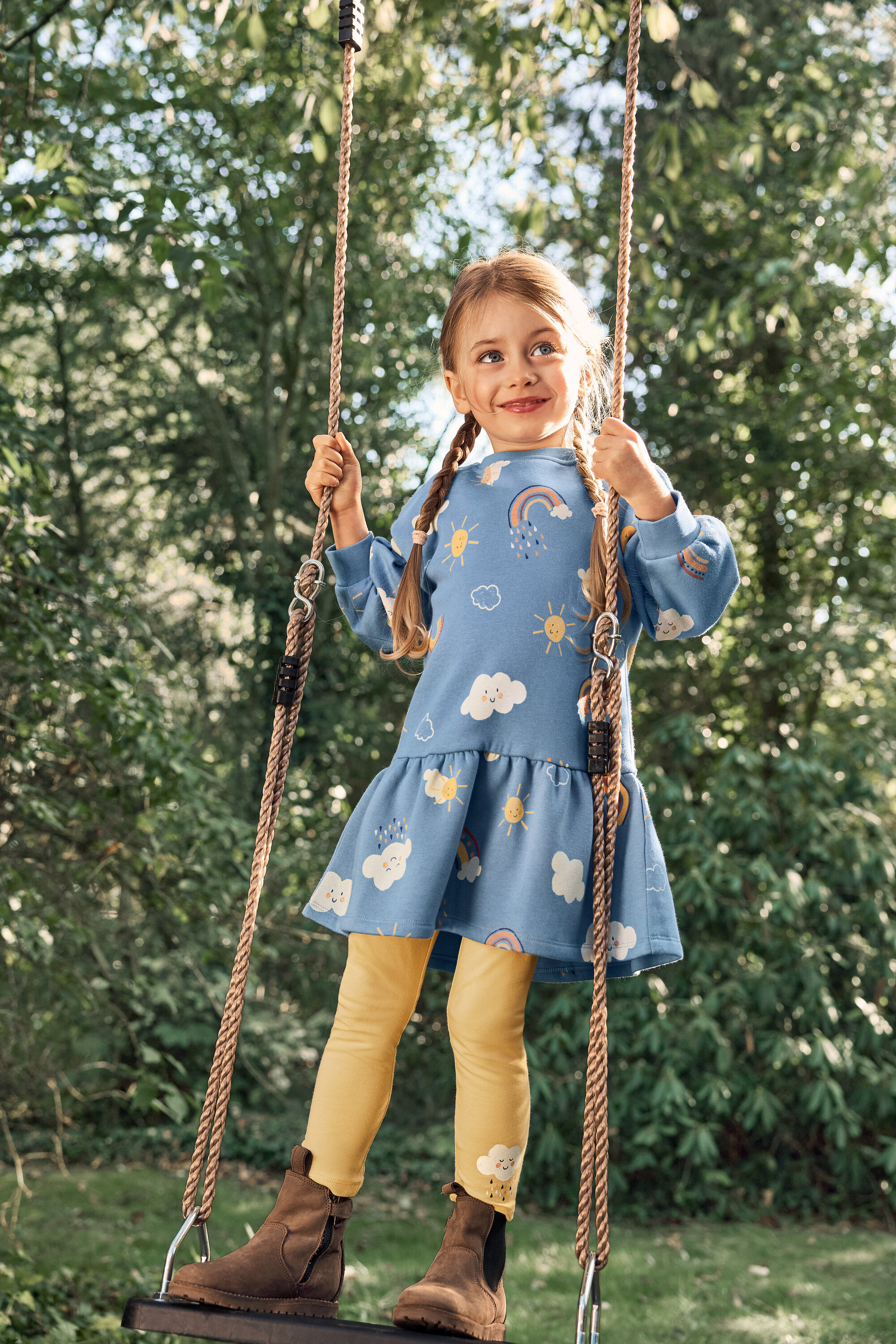 Egy copfos kislány hintázik egy hintán, kék ruhát visel nap-, felhő- és szivárványmintával, sárga leggingset és barna csizmát.