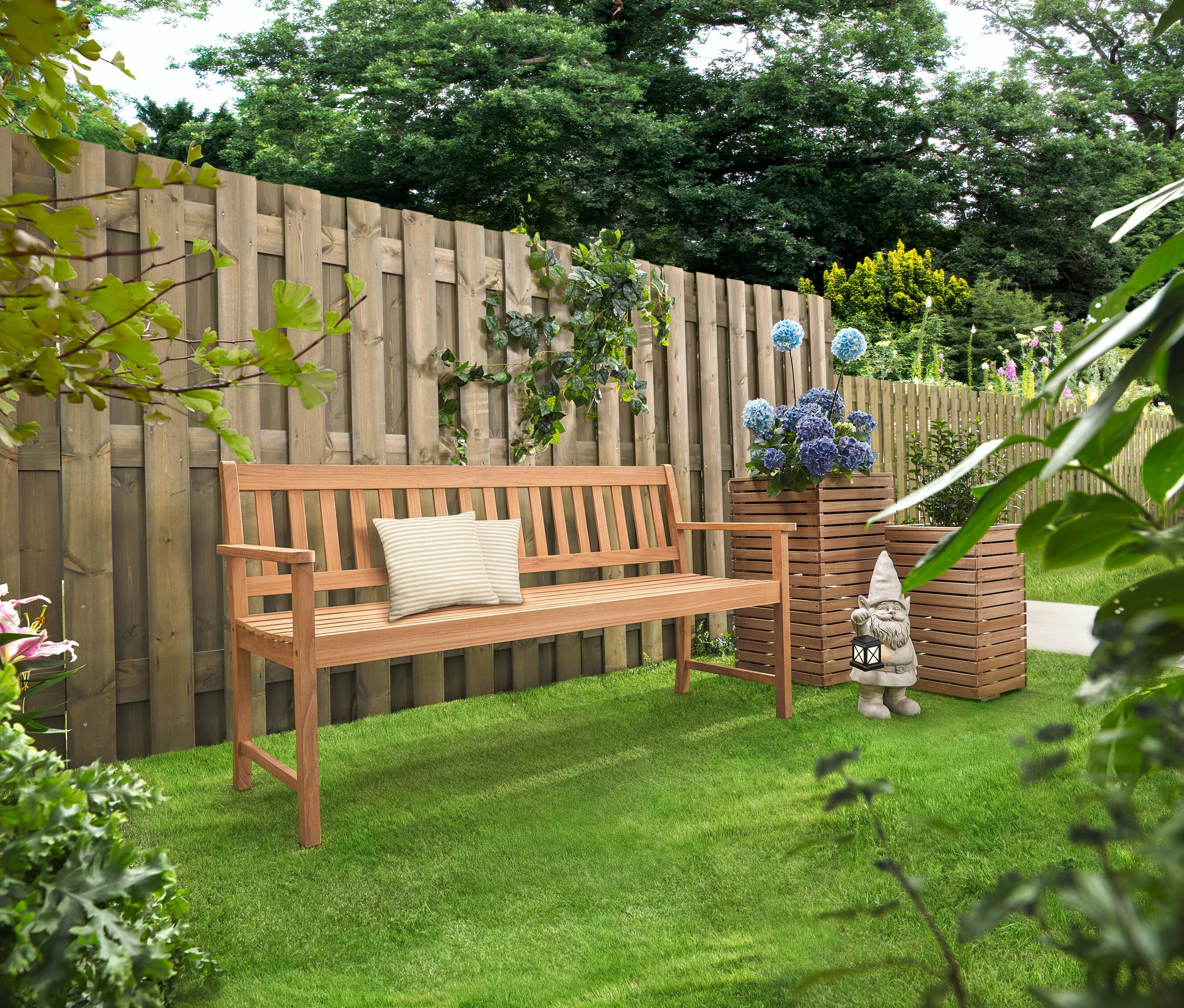 Egy fa pad párnákkal áll a füvön. Mögötte két »Tinus« virágoszlop áll kék virágokkal.