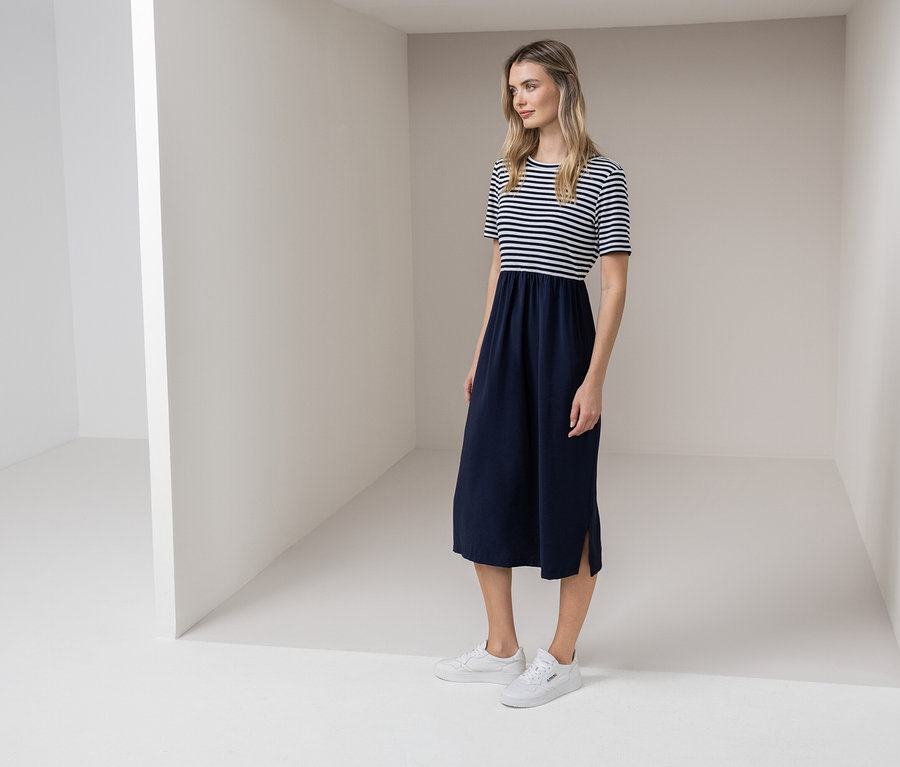 Egy nő áll egy fehér szobában, kék-fehér csíkos ruhát és fehér tornacipőt visel.