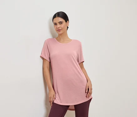 Rózsaszín pólót és leggingset viselő nő egy fehér falnak támaszkodik.
