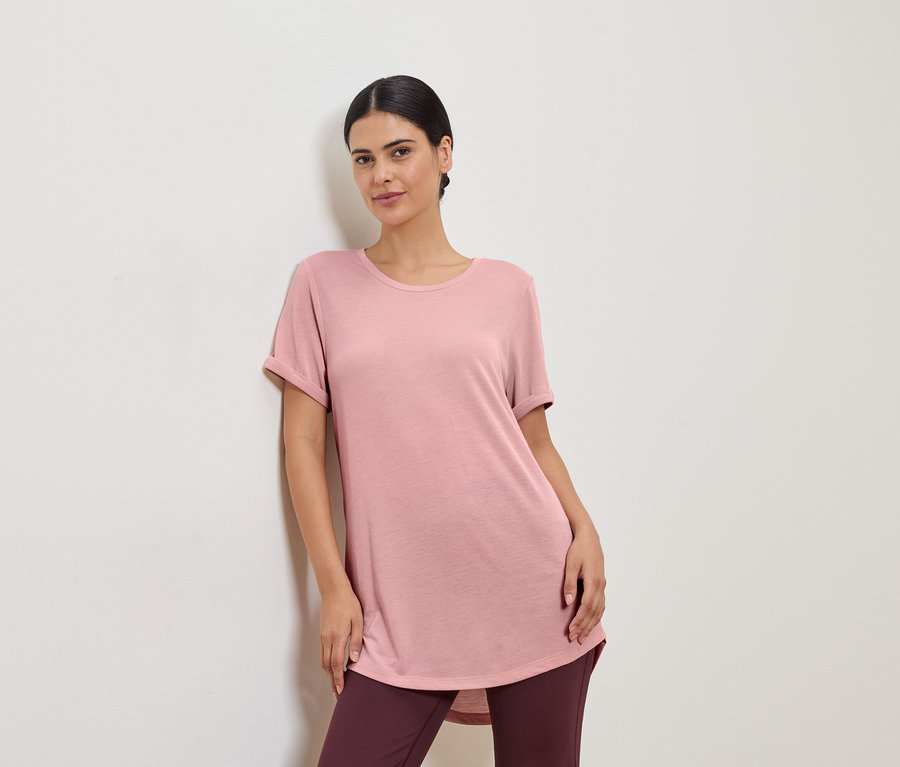 Rózsaszín pólót és leggingset viselő nő egy fehér falnak támaszkodik.