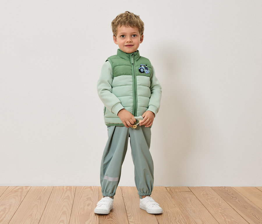 Egy fiú áll a fal előtt világoszöld kabátban, világoszöld nadrágban és fehér tornacipőben.