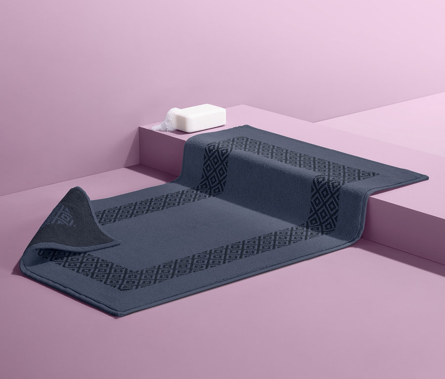 Egy kék fürdőszoba szőnyeg geometrikus mintával rózsaszín alapon. Rajta egy fehér szappan.