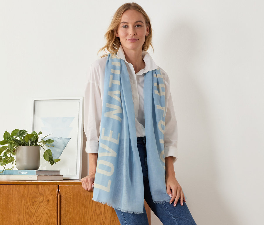 Nő támaszkodik egy szekrénynek, világoskék, feliratos női szövetsálat visel.