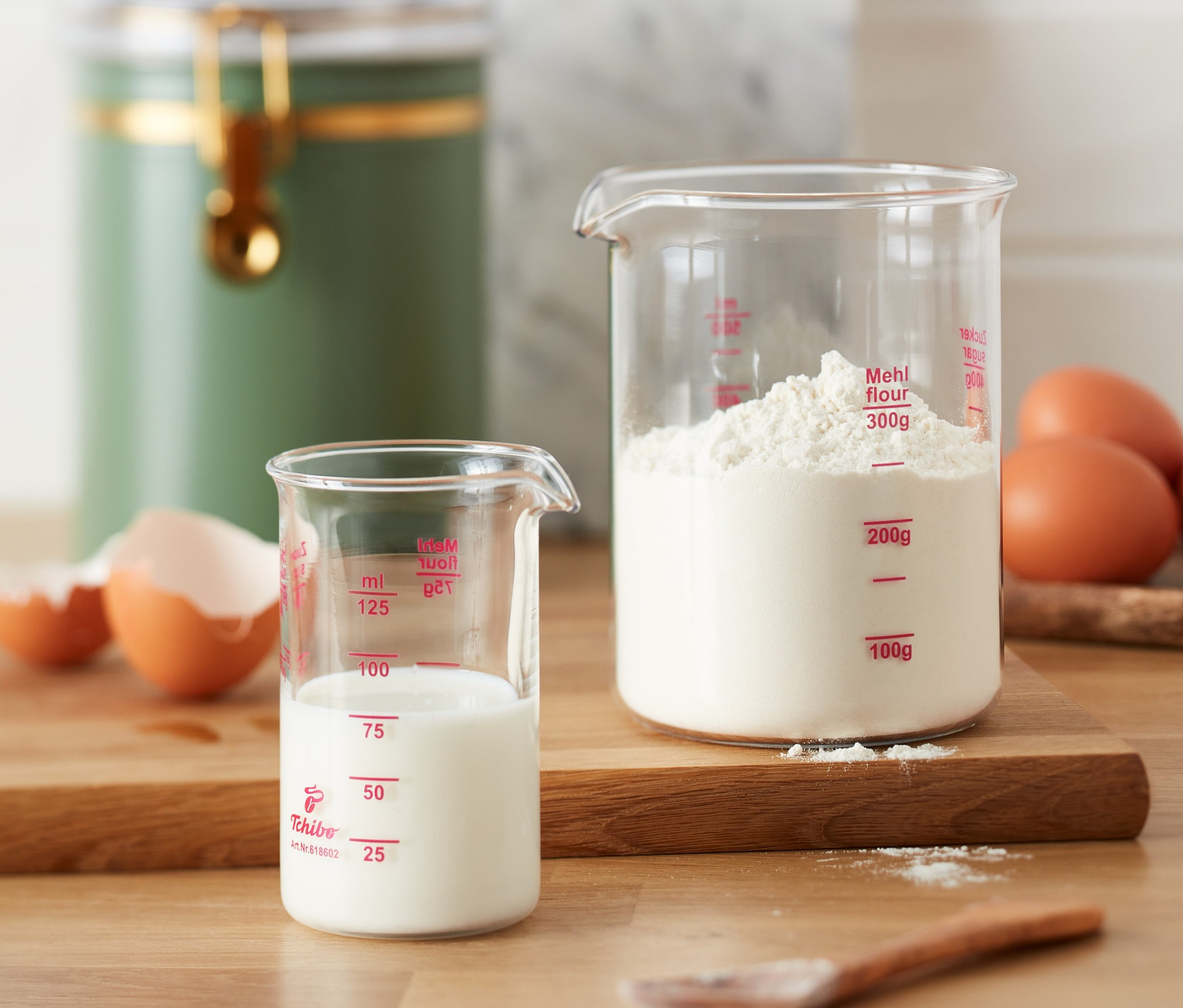Két mérőpohár, az egyikben tej, a másikban liszt van, a faasztalon tojások mellett.