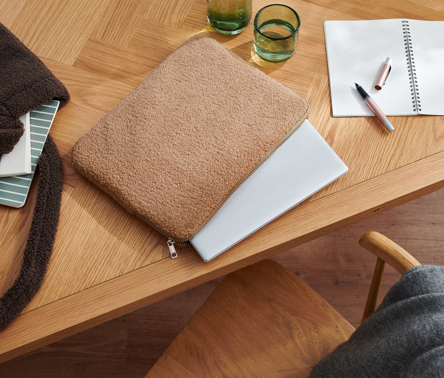 Egy fa asztalon egy laptoptáska, teddyszőrmés hatás, egy notebook, egy nyitott jegyzettömb, egy toll és poharak találhatók.