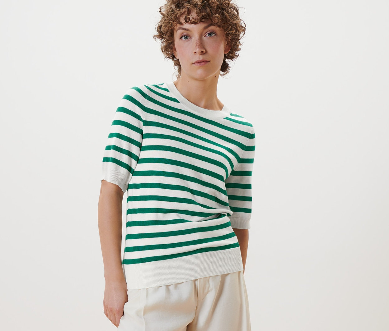 Nő pózol egy finom kötésű csíkos pulóverben, fehér nadrágban és fehér cipőben.