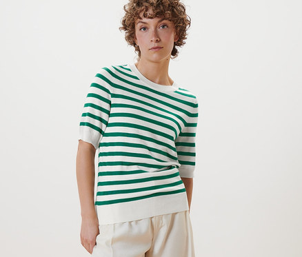 Nő pózol egy finom kötésű csíkos pulóverben, fehér nadrágban és fehér cipőben.