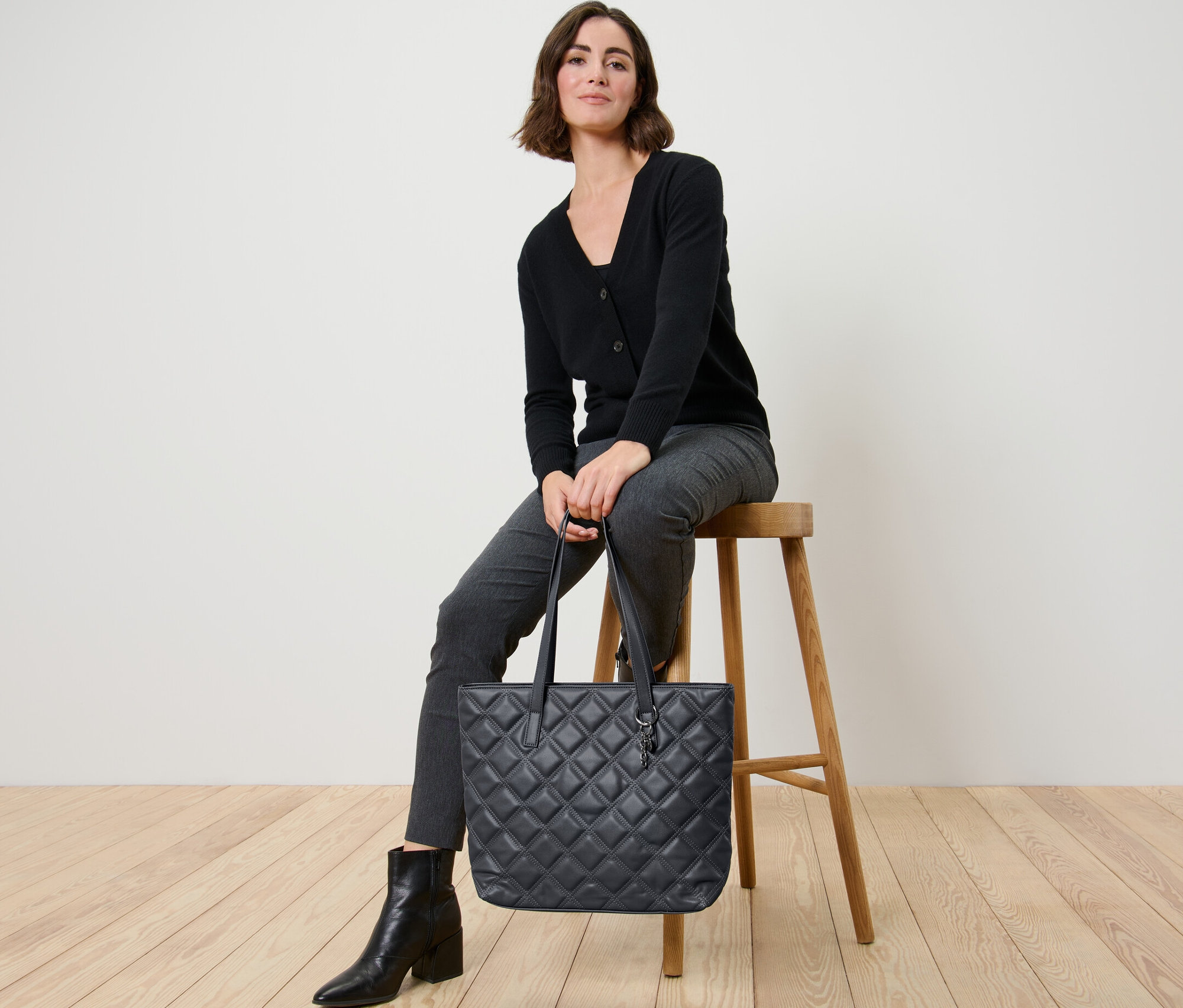 Nő ül a széken és egy sötétszürke, műbőr, steppelt shopper táskát tart.