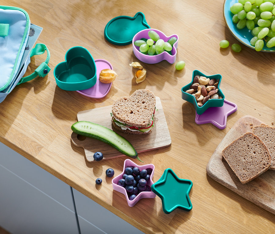 Egy fa asztalon egy uzsonna található. Szív alakú szendvics, uborka, áfonya egy csillag alakú edényben, diók, szőlő egy hal alakú edényben és kenyér.