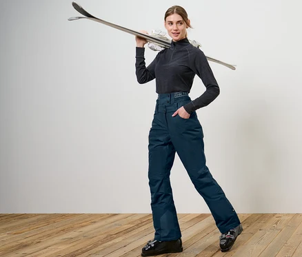 Nő pózol tengerészkék sí- és snowboardnadrágban, a vállán síléccel.