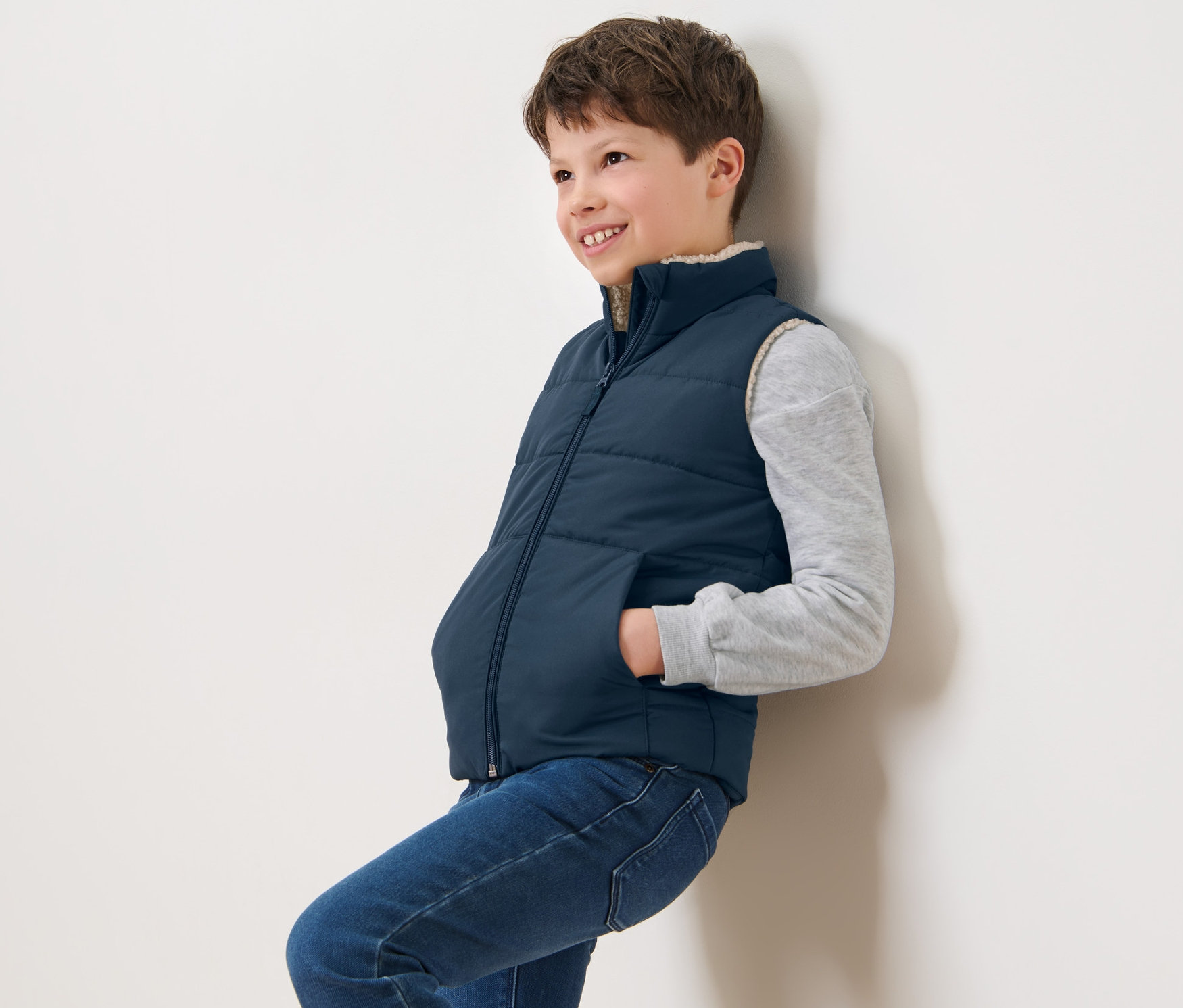 Egy falnak támaszkodó fiú sötétkék gyerek steppelt mellényt visel, plüssbéléssel.