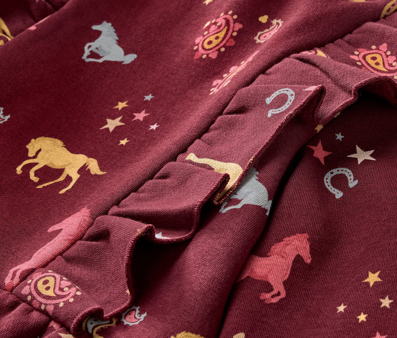 Egy bordó színű, fodros, lovas, csillagos és patkómintás gyerekruha részletének közeli képe.