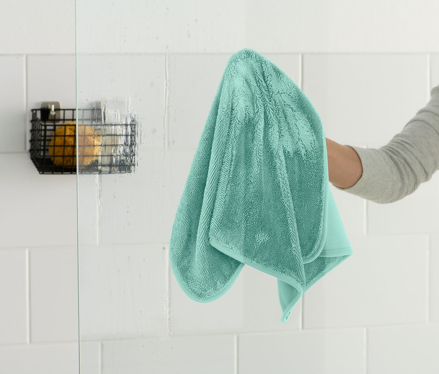 Egy személy töröl egy üveg zuhanyfalat egy türkiz törülközővel. A sarokban egy fekete drótkosár látható.
