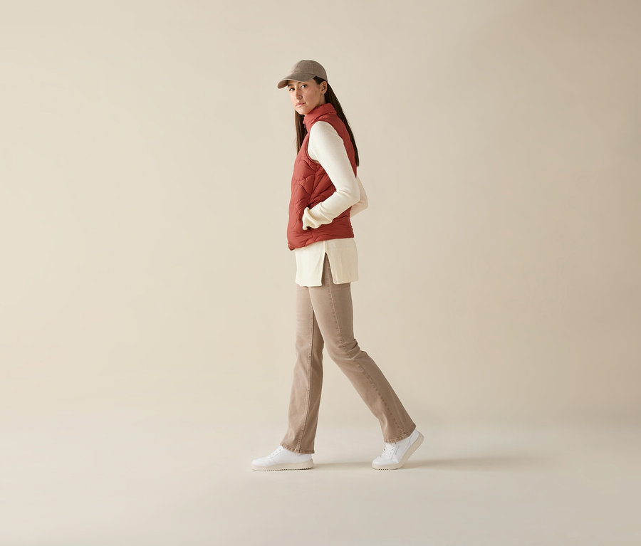 Nő sétál, steppelt mellényt, hosszú ujjú felsőt, nadrágot és baseballsapkát visel.