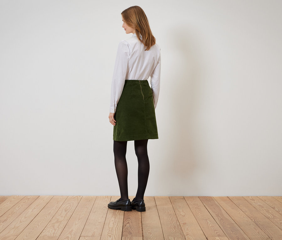 Egy nő háttal áll, zöld kordbársony szoknyát, fehér inget, fekete harisnyát és fekete cipőt visel.