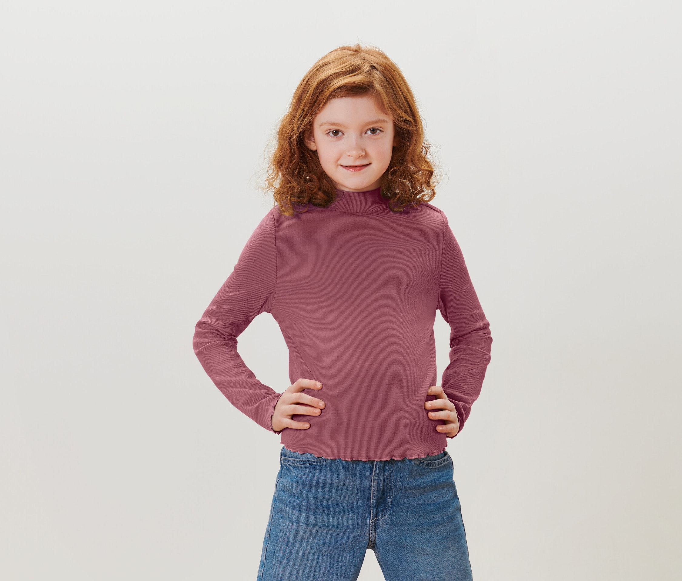 Egy vörös, göndör hajú lány csípőre tett kézzel áll. Lila hosszú ujjú inget és kék farmert visel.