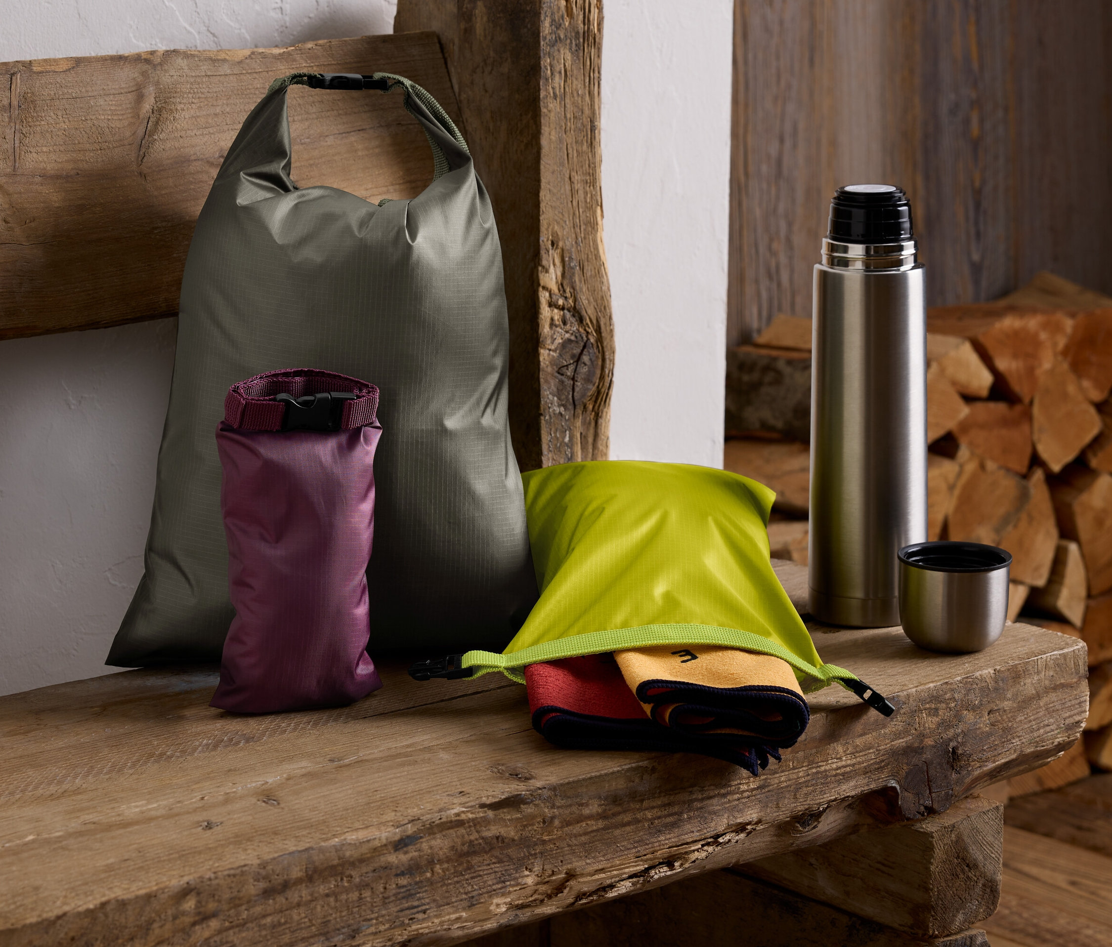 Egy fa padon egy zöld zsák, egy lila zsák, egy zöld zsák törölközőkkel, egy termosz és egy rakat fa van.