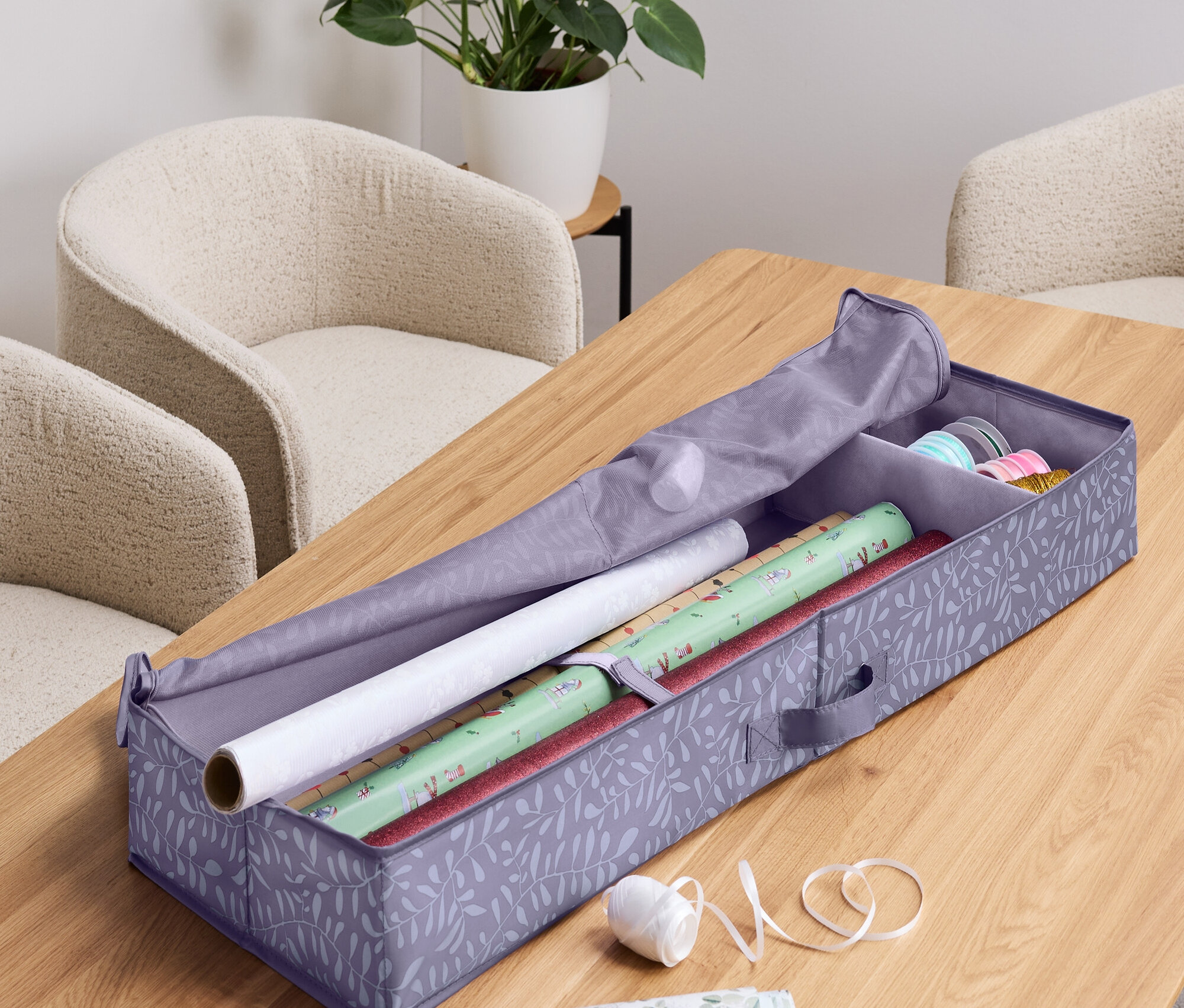 Nyitott tárolódoboz csomagolópapírhoz papírtekercsekkel, szalaggal, ollóval és egy növénnyel a háttérben.