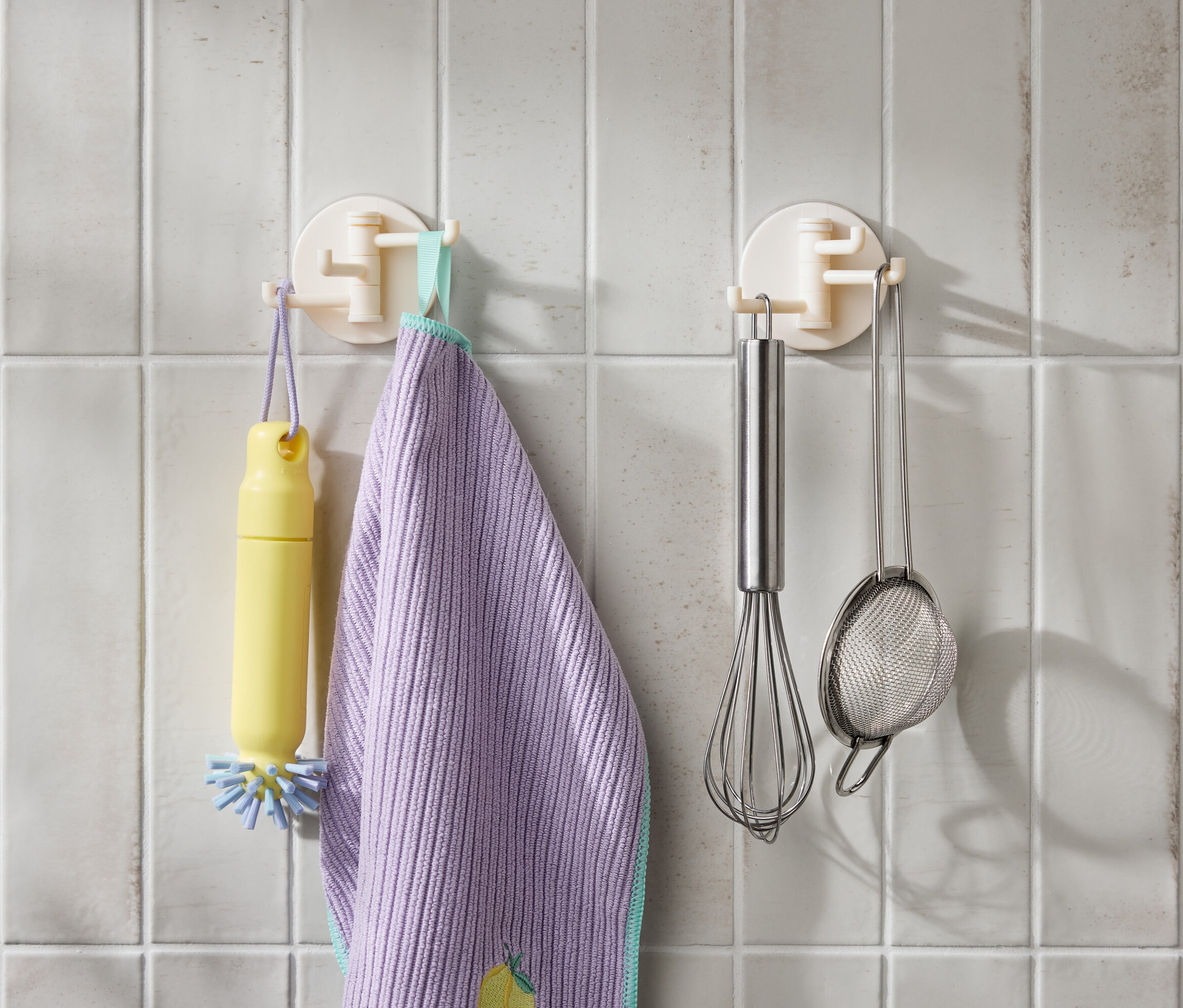 Fehér csempén lóg egy sárga kefe, egy lila törlőkendő, egy rozsdamentes habverő és egy szűrő.