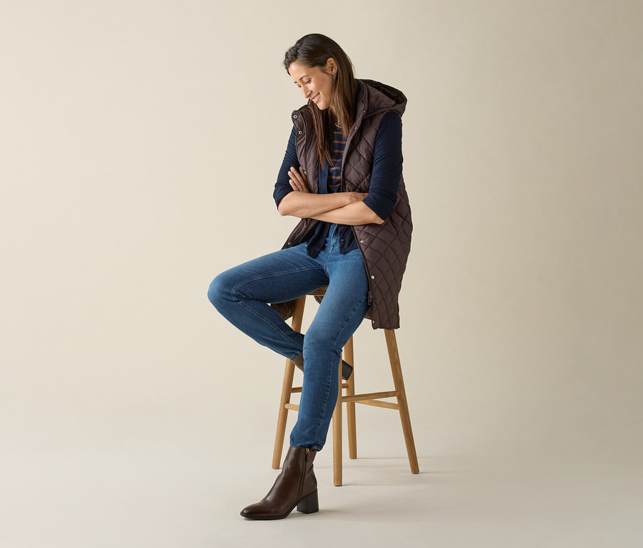 Egy nő ül egy széken keresztbe tett karokkal, és egy hosszú, sötétbarna, steppelt mellényt visel kapucnival.