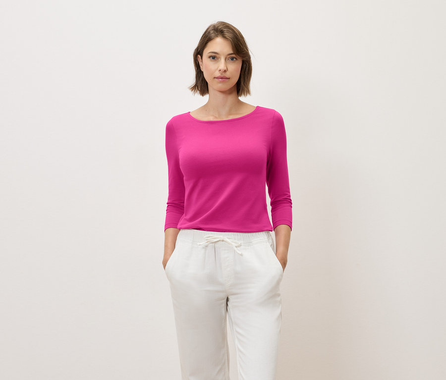 Barna hajú nő rózsaszín, háromnegyedes ujjú felsőben és fehér nadrágban pózol.